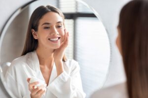 Woman smiling in mirror while she applies moisturizer to help prevent winter dryness